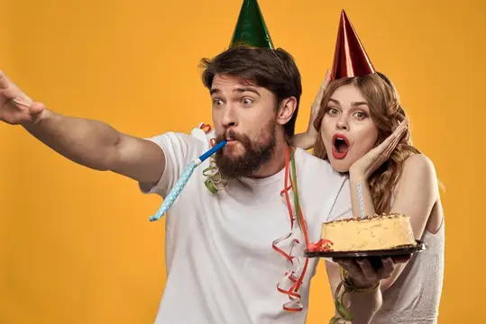 Мужчина и женщина в колпаках дуют в дудки с тортом