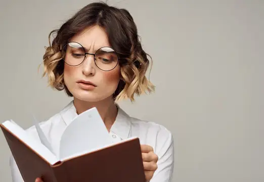 Молодая женщина в очках сосредоточенно читает книгу на сером фоне