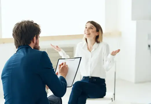 Улыбчивая женщина в белой рубашке жестикулирует на встрече с мужчиной с планшетом