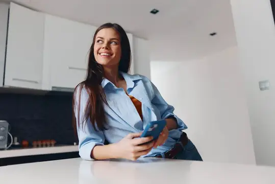 Счастливая женщина стоит на кухне с мобильным телефоном и улыбается