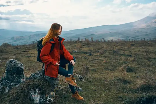Рыжеволосая девушка в красной куртке сидит на камне в горах
