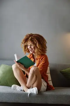 Кудрявая девушка в оранжевой рубашке смеётся, читая книгу на диване