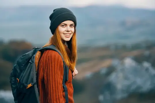 Рыжеволосая девушка в шапке с рюкзаком улыбается на фоне гор