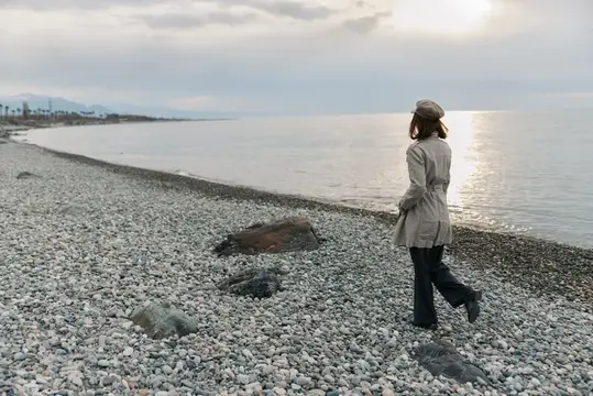 Девушка в плаще идёт по галечному пляжу у моря