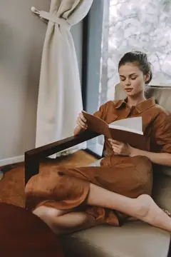 Молодая женщина в коричневом платье читает книгу в кресле у окна