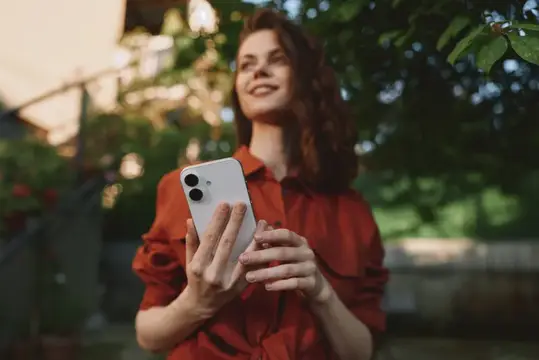 Улыбающаяся девушка в красной рубашке держит смартфон на улице