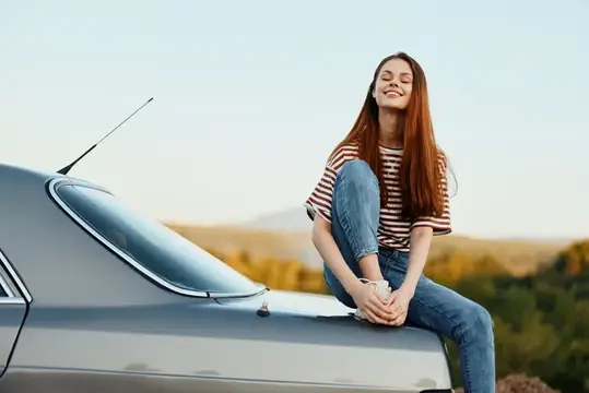 Женщина-водитель сидит на багажнике автомобиля и улыбается, любуясь красивым видом осенней природы и гор