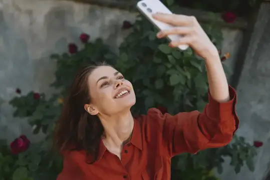 Улыбающаяся темноволосая женщина делает селфи на фоне розовых кустов
