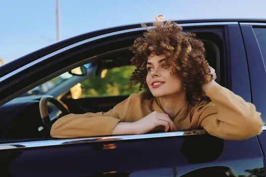 Кудрявая девушка в бежевом топе выглядывает из окна автомобиля