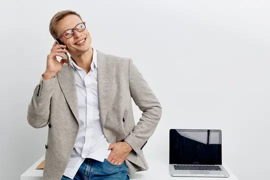 Счастливая женщина пользуется мобильным телефоном в офисной обстановке с ноутбуком на столе, повседневная деловая одежда, жизнерадостное и уверенное настроение, очки, светло-серый блейзер, белая…