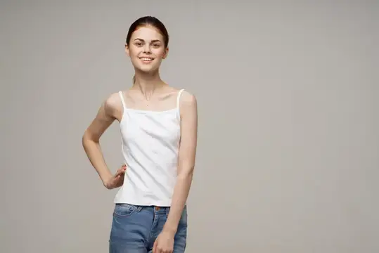 Улыбающаяся девушка в белой майке и джинсах на сером фоне