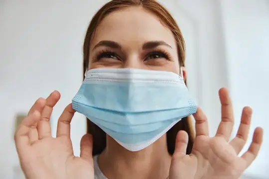Улыбающаяся девушка поправляет голубую медицинскую маску