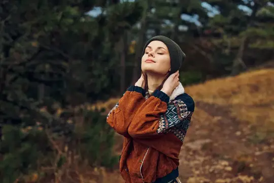 Молодая женщина в шапке и куртке с закрытыми глазами обнимает себя в осеннем лесу