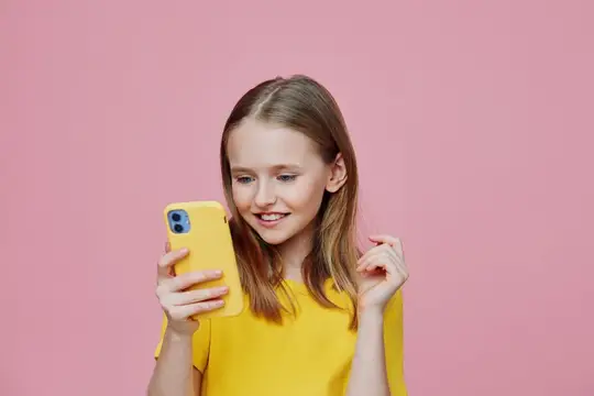 Светловолосая девочка смотрит в жёлтый смартфон на розовом фоне