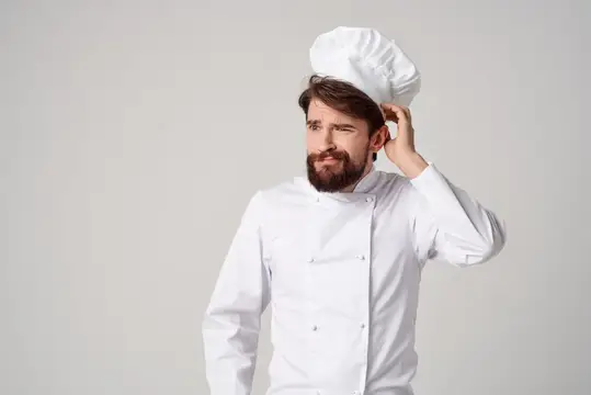 Задумчивый бородатый повар в белом колпаке почесывает голову