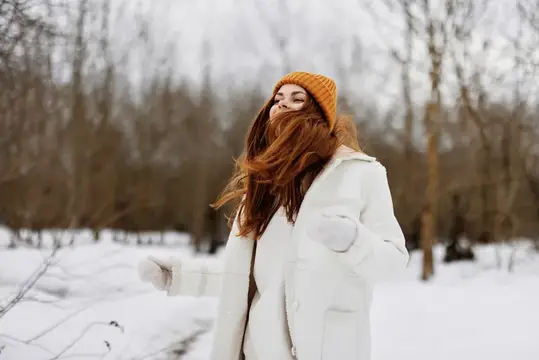 Рыжеволосая девушка в оранжевой шапке весело кружится в зимнем лесу