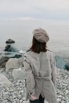 Вид со спины на женщину в клетчатом пальто и берете, смотрящую на море с каменистого пляжа