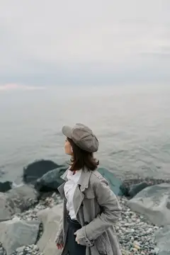 Молодая женщина в кепке и плаще смотрит на море с каменистого берега