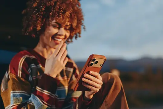 Рыжеволосая женщина смеётся, глядя в смартфон на закате