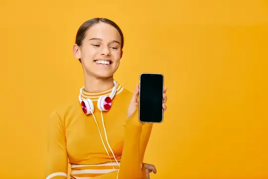 Улыбчивая девушка с наушниками показывает смартфон на жёлтом фоне