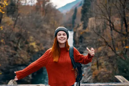 Улыбающаяся рыжеволосая девушка в оранжевом свитере на фоне осеннего водопада