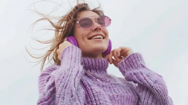 Счастливая женщина в фиолетовом свитере и розовых наушниках-повязке слушает музыку с улыбкой