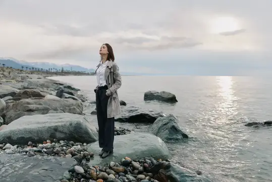 Молодая женщина в плаще стоит на камнях у моря с закрытыми глазами