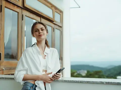 Молодая женщина со смартфоном стоит у окна с видом на горы