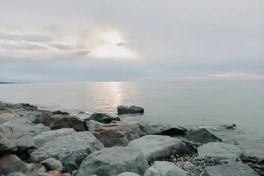 Спокойное море с каменистым берегом под облачным небом, мягкий солнечный свет отражается на поверхности воды