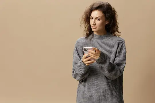 Кудрявая девушка в сером свитере смотрит в смартфон на бежевом фоне