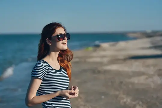 Женщина в очках и полосатой футболке гуляет по пляжу летом