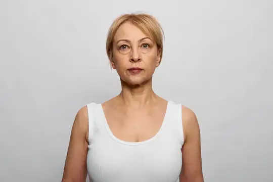Светловолосая женщина средних лет в белой майке на сером фоне