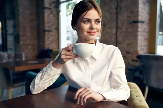 Улыбающаяся девушка в белой рубашке пьёт кофе в кафе