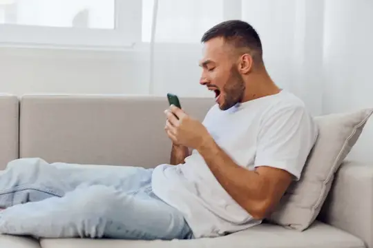Это изображение запечатлело удивленного мужчину, использующего смартфон в уютной гостиной, что символизирует эмоциональное влияние технологий на личную жизнь