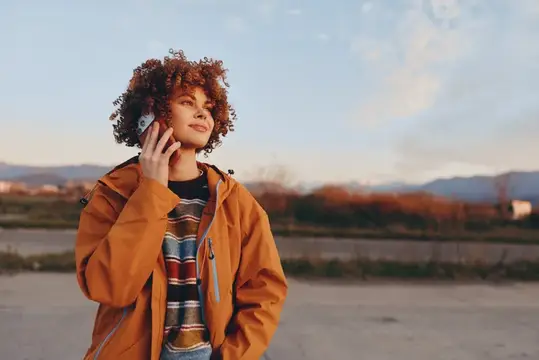 Рыжеволосая девушка с кудрями улыбается, слушая телефон на улице