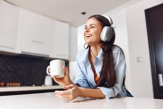 Улыбающаяся темноволосая девушка в наушниках с кружкой на кухне