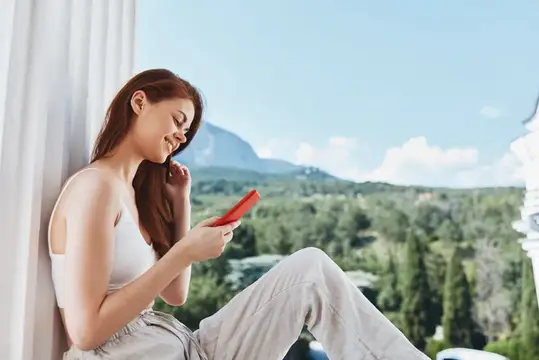 Улыбающаяся девушка с красным телефоном у окна с видом на горы