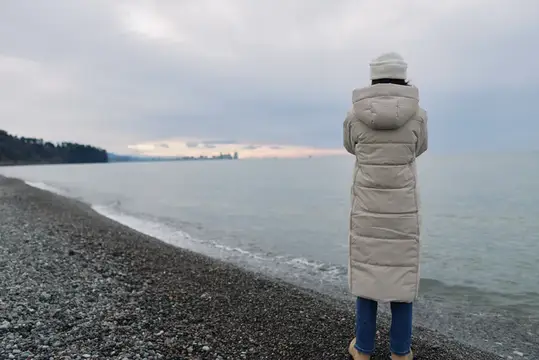 Женщина в длинном пуховике смотрит на море с галечного пляжа