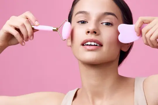 Молодая женщина с массажным роллером и скребком для лица на розовом фоне