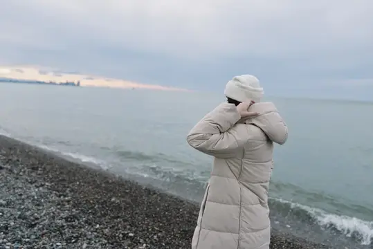 Женщина в светлом пуховике стоит на галечном берегу у моря