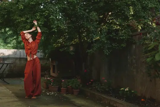 Энергичная сцена из жизни женщины: развевающийся ржавый комбинезон, танцует в саду рядом с горшками и кирпичной стеной, искреннее и приподнятое настроение, естественный солнечный свет, передача…