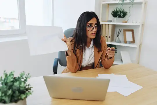 Бизнес-леди работает в офисе за столом с ноутбуком, испытывая гнев и участвуя в споре, обсуждая бизнес-процессы онлайн по видеосвязи, демонстрируя документы и отчеты