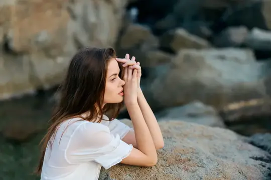 Задумчивая темноволосая девушка в белой блузке у скал