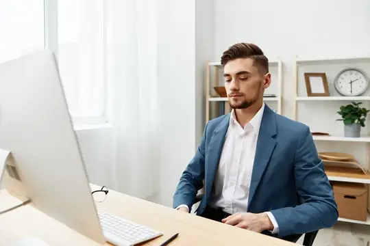 Молодой мужчина в синем пиджаке работает за компьютером в светлом офисе