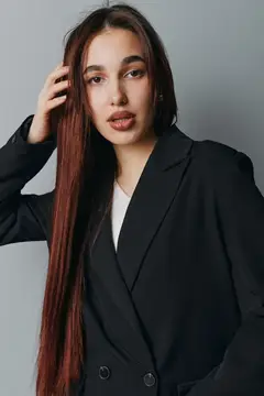 Модель женского пола с прямыми длинными волосами, одетая в черную куртку и белый топ, стоит перед однотонной серой стеной