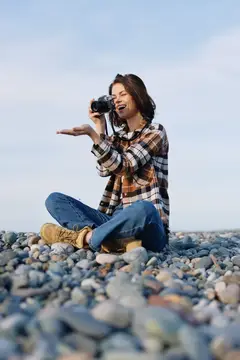 Молодая женщина с фотокамерой сидит на каменистом берегу и улыбается