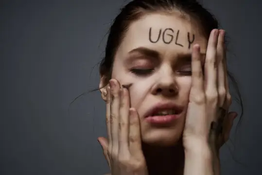 Молодая женщина с надписью UGLY на лбу закрывает лицо руками