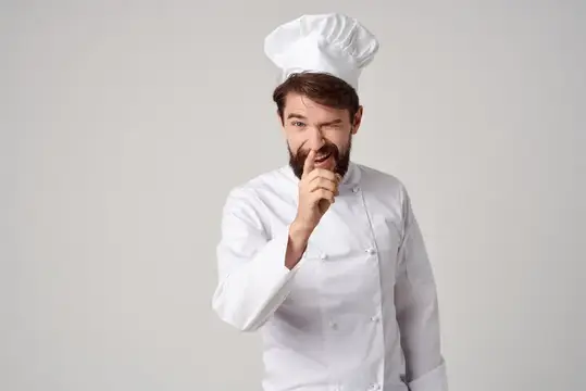 Бородатый повар в белом колпаке прикладывает палец к губам