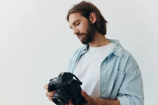 Фотограф-хипстер в студии на белом фоне смотрит на экран камеры и настраивает её для фотосъёмки