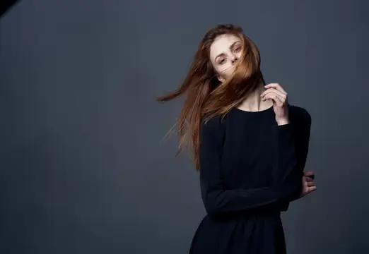 Рыжеволосая девушка в чёрном платье отводит волосы от лица на сером фоне